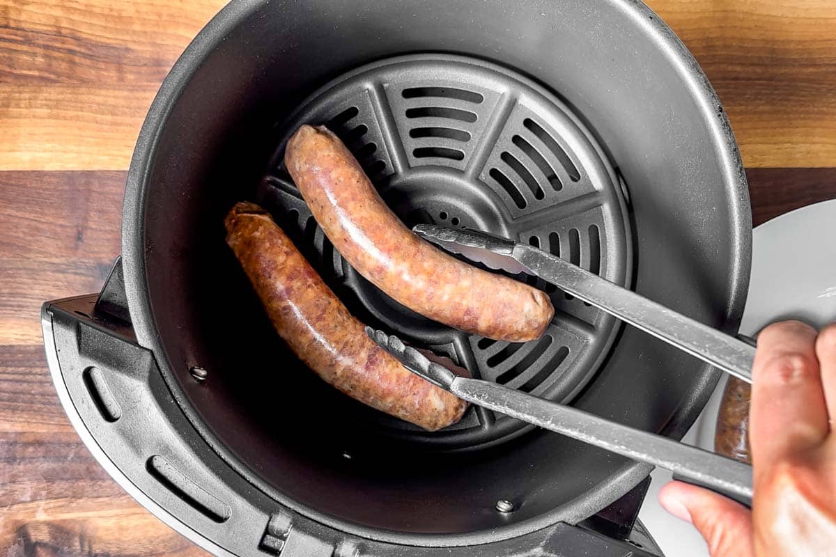 Placing the Italian sausages into an air fryer with a pair of tongs.