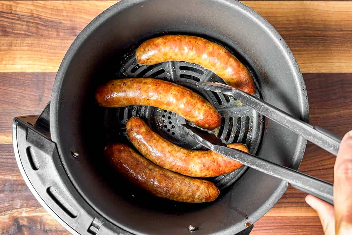 Finished browned Italian sausages being removed from the air fryer with a pair of tongs.