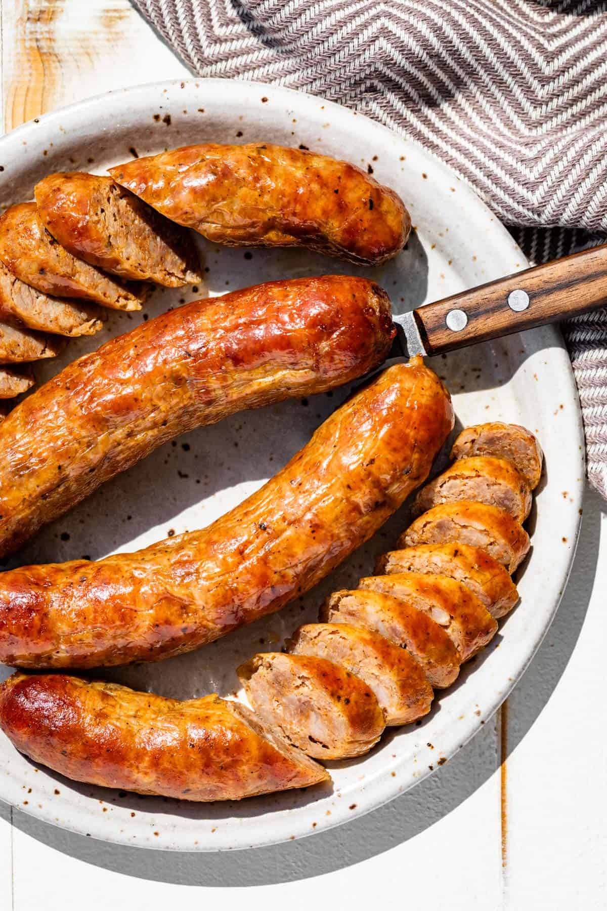 Four Italian sausages browned and partially sliced on a pottery plate with a wood handled knife on the side.