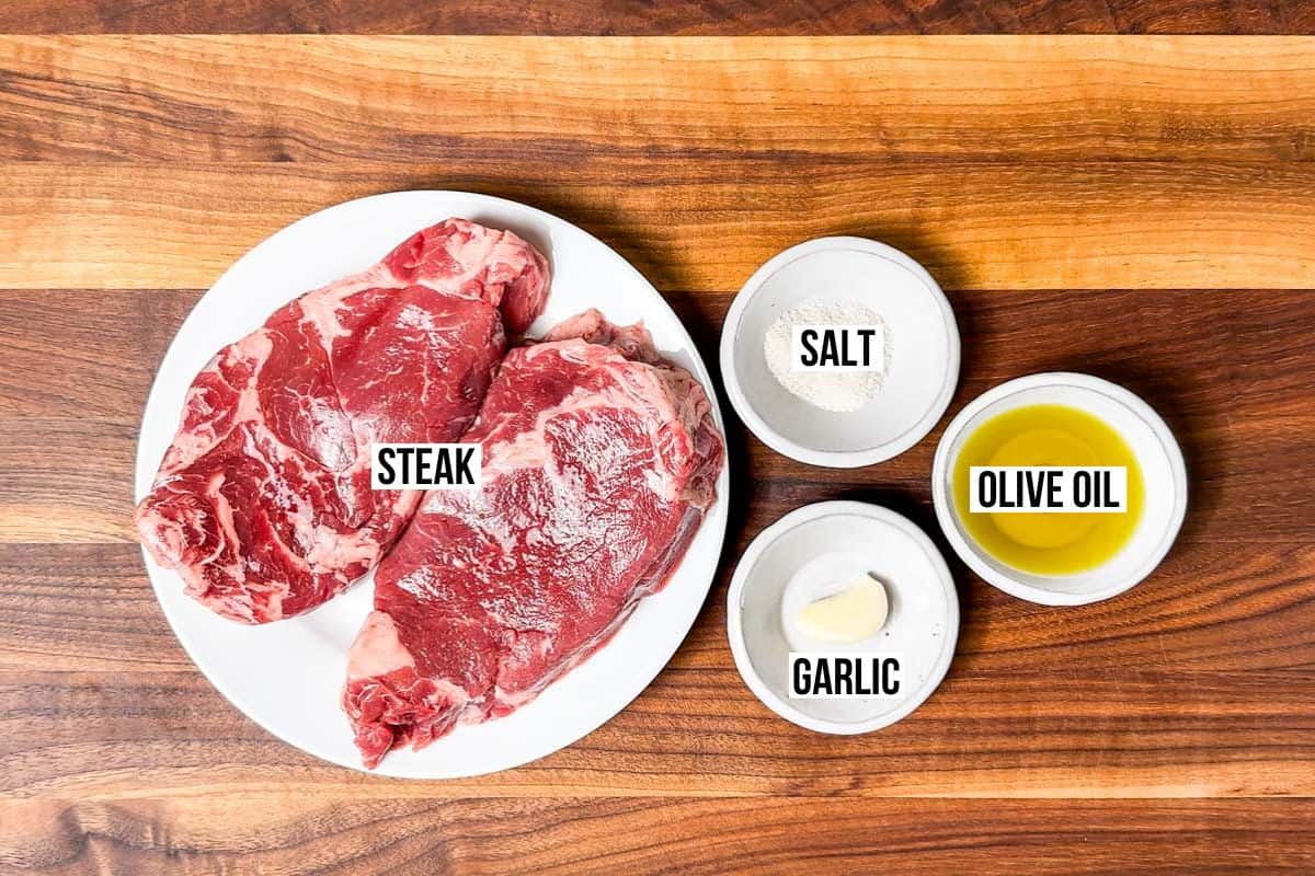 Ribeye steaks, sea salt, garlic, and olive oil in containers on a wood cutting board.