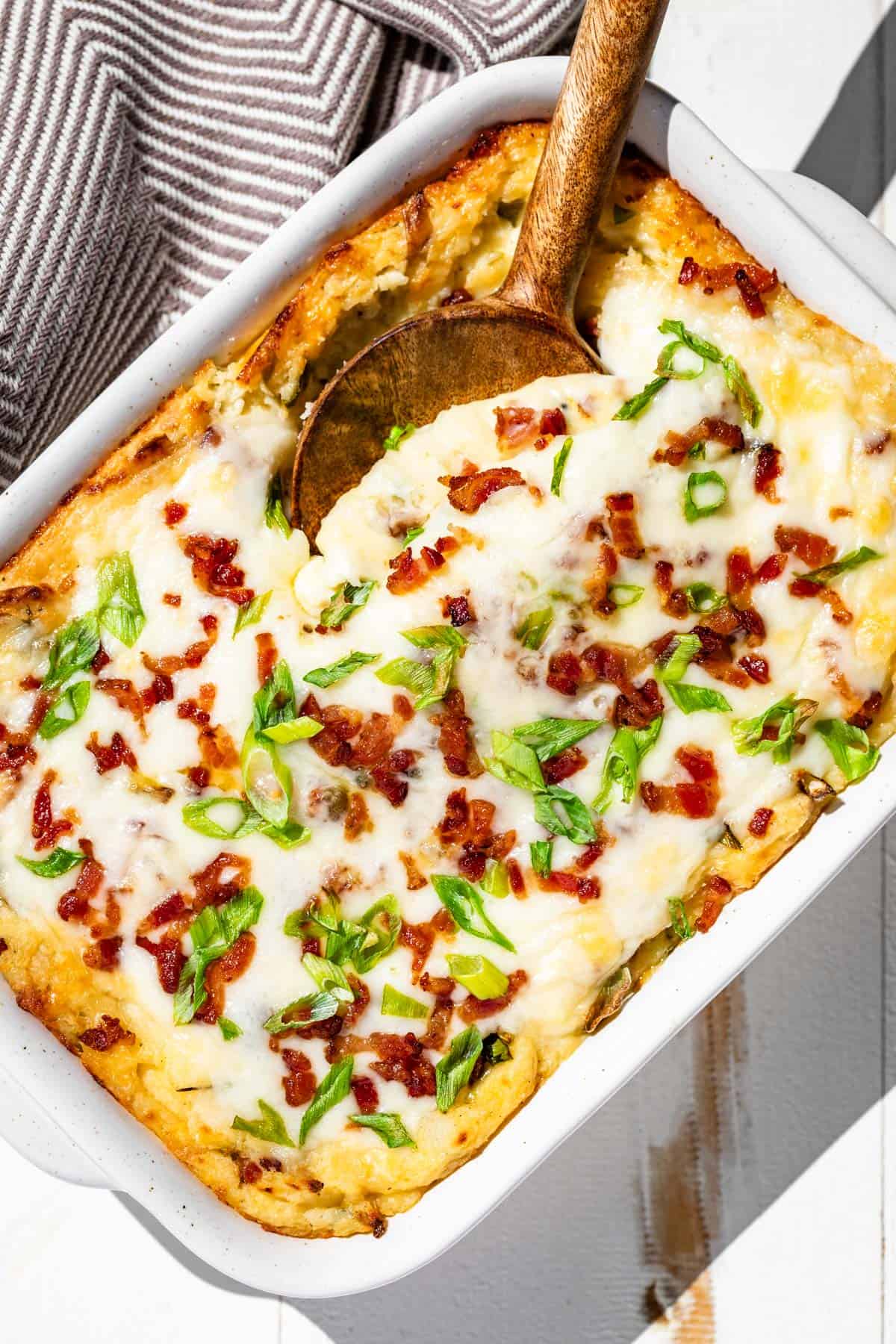 A pottery baking dish with baked cheesy mashed potatoes topped with cheese, bacon bits, and sliced green onion.