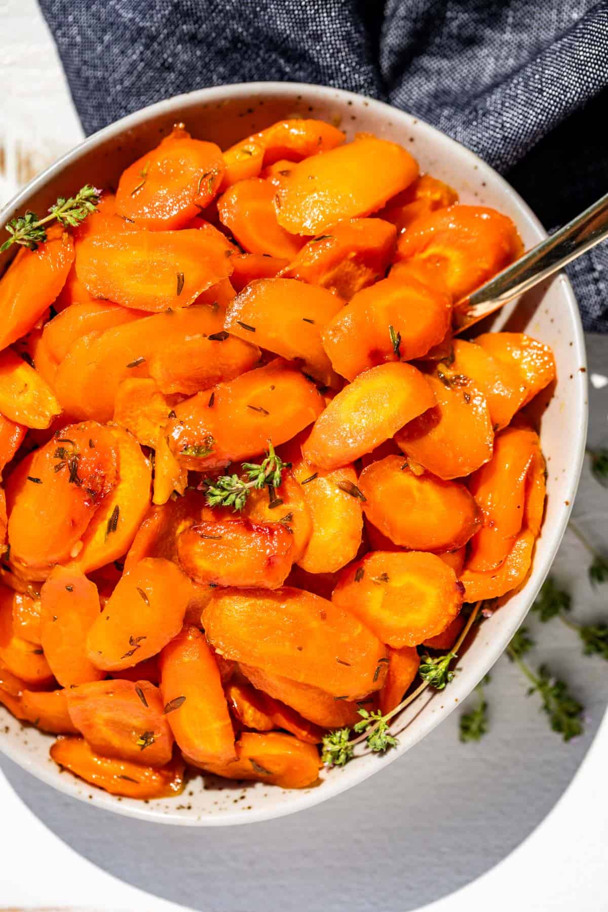 A taupe pottery bowl filled with sliced honey glazed carrots with a gold spoon on the side and garnished with a sprig of thyme.