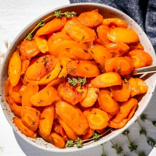 Finished honey glazed carrots in a taupe colored pottery bowl with a gold spoon in the bowl and a blue linen on the side.