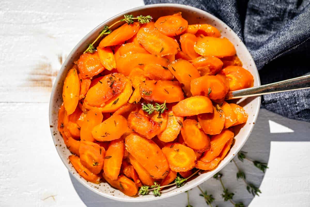 Finished honey glazed carrots in a taupe colored pottery bowl with a gold spoon in the bowl and a blue linen on the side.