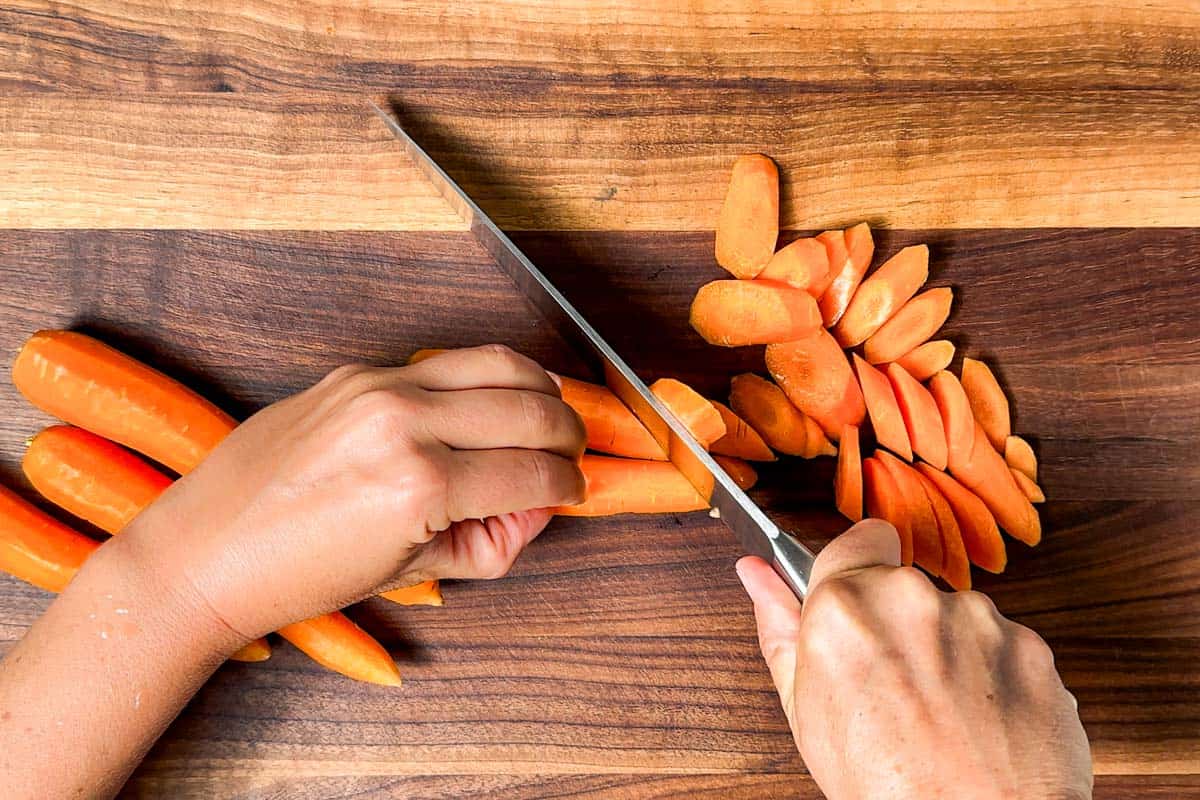 Slicing carrots with a large chefs knife on a wood cutting board.