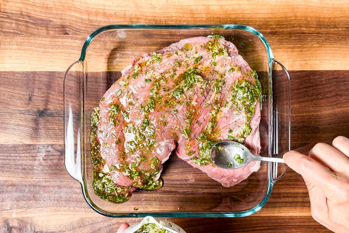 Coating one side of the tri tip with marinade in a glass container on a wood cutting board.