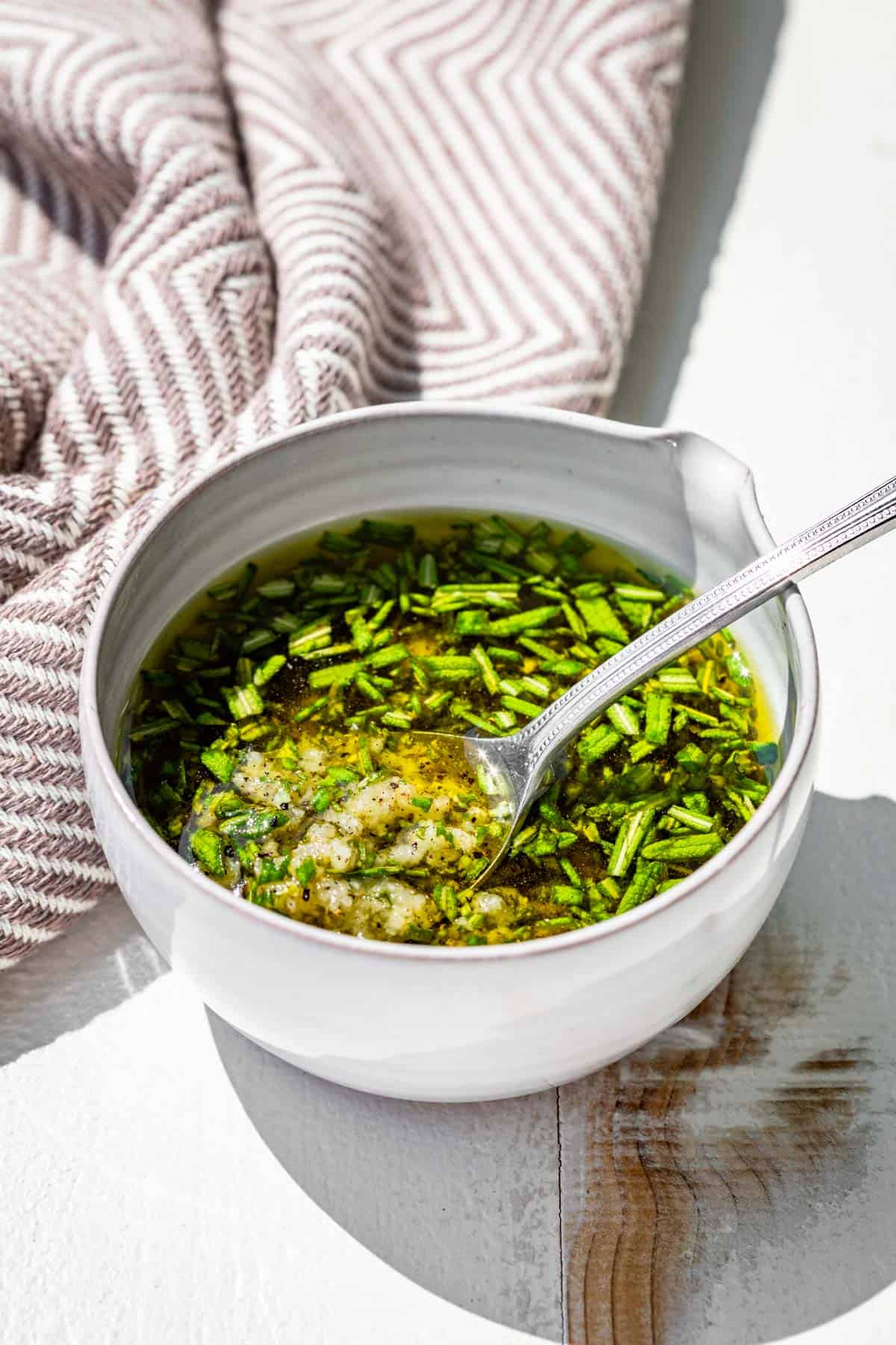 Mixing together the rosemary, olive oil, minced garlic, sea salt, and pepper in a small pottery bowl with a spout.