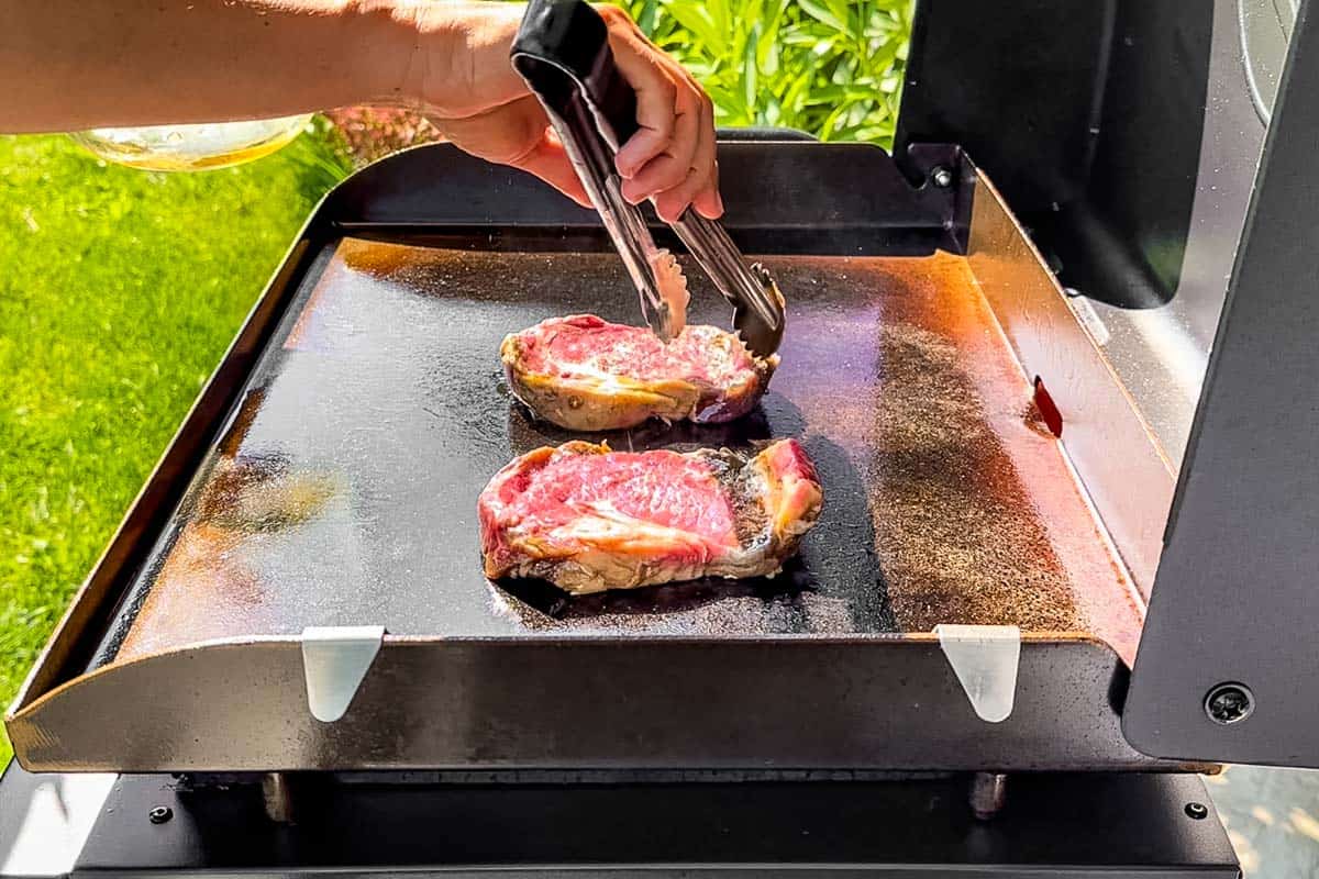 Placing the marinated steak on a Blackstone griddle outside with metal tongs and green grass in the background.