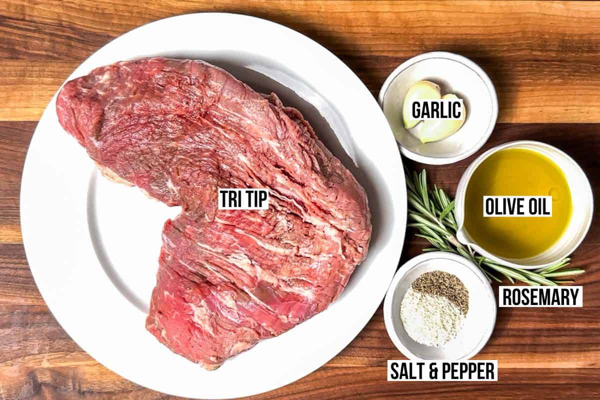 A tri tip on a large white plate with bowls of garlic, olive oil, sea salt and pepper on the side with sprigs of rosemary.