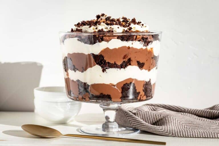 A large clear glass trifle bowl filled with cubes of chocolate cake, chocolate pudding, and whipped cream in layers with bowls and a serving spoon on the side.