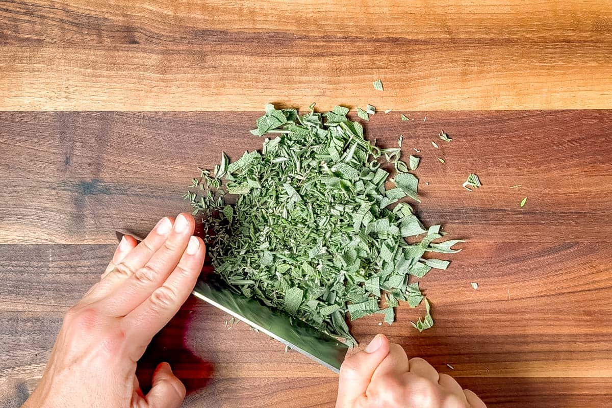 Chopping the rosemary, sage, and thyme with a large knife on a wood cutting board.