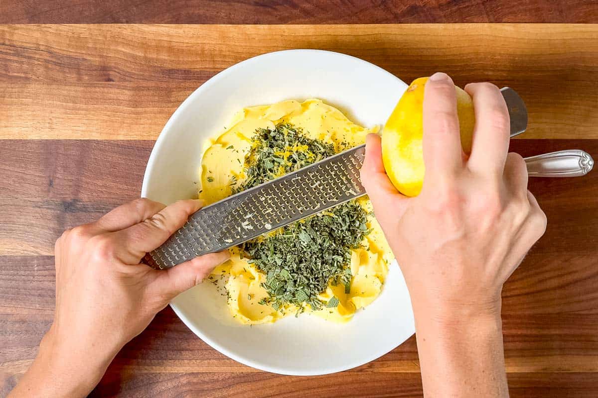 Zesting a lemon over the herbs and butter in a large white bowl.