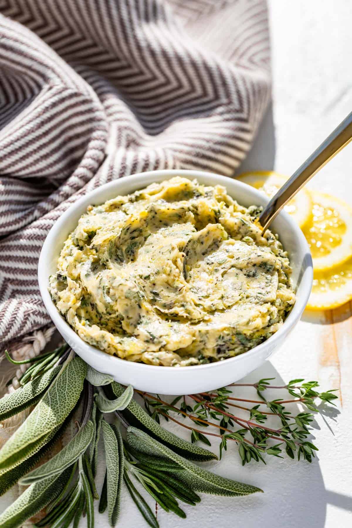 Herb butter in a white bowl with a brown linen in the background and sprigs of fresh herbs and slices lemon on the side of the bowl.