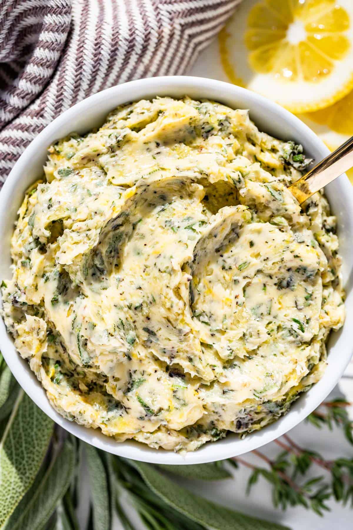 Herb butter in a white bowl with swirls on top and fresh herbs and lemon slices on the side.