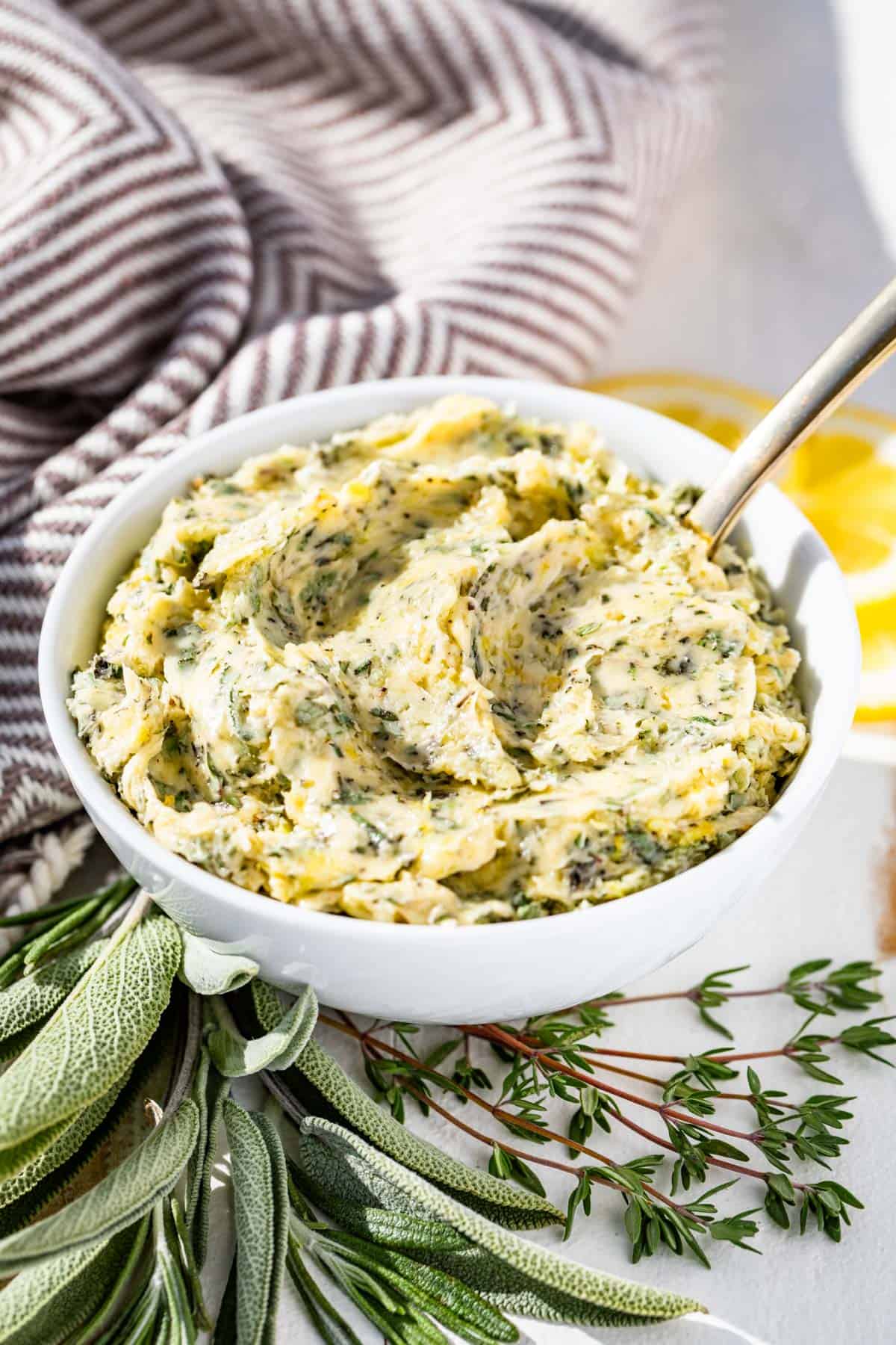 A white bowl filled with herb butter with a silver spoon in it with sprigs of sage, rosemary, and thyme around it.