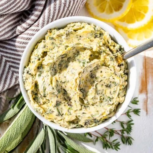 Finished herb butter in a white bowl with a silver spoon with sprigs of rosemary, sage, and thyme and lemon slices on the side.