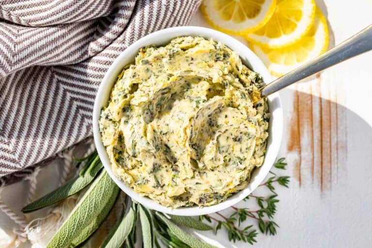 Finished herb butter in a white bowl with a silver spoon with sprigs of rosemary, sage, and thyme and lemon slices on the side.