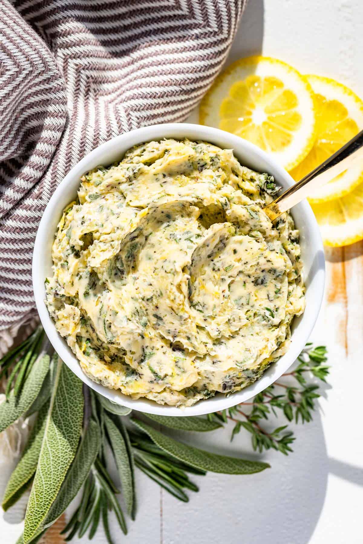 A white bowl filled with herb butter surrounded by sage, rosemary, and thyme with lemon slices on the side.