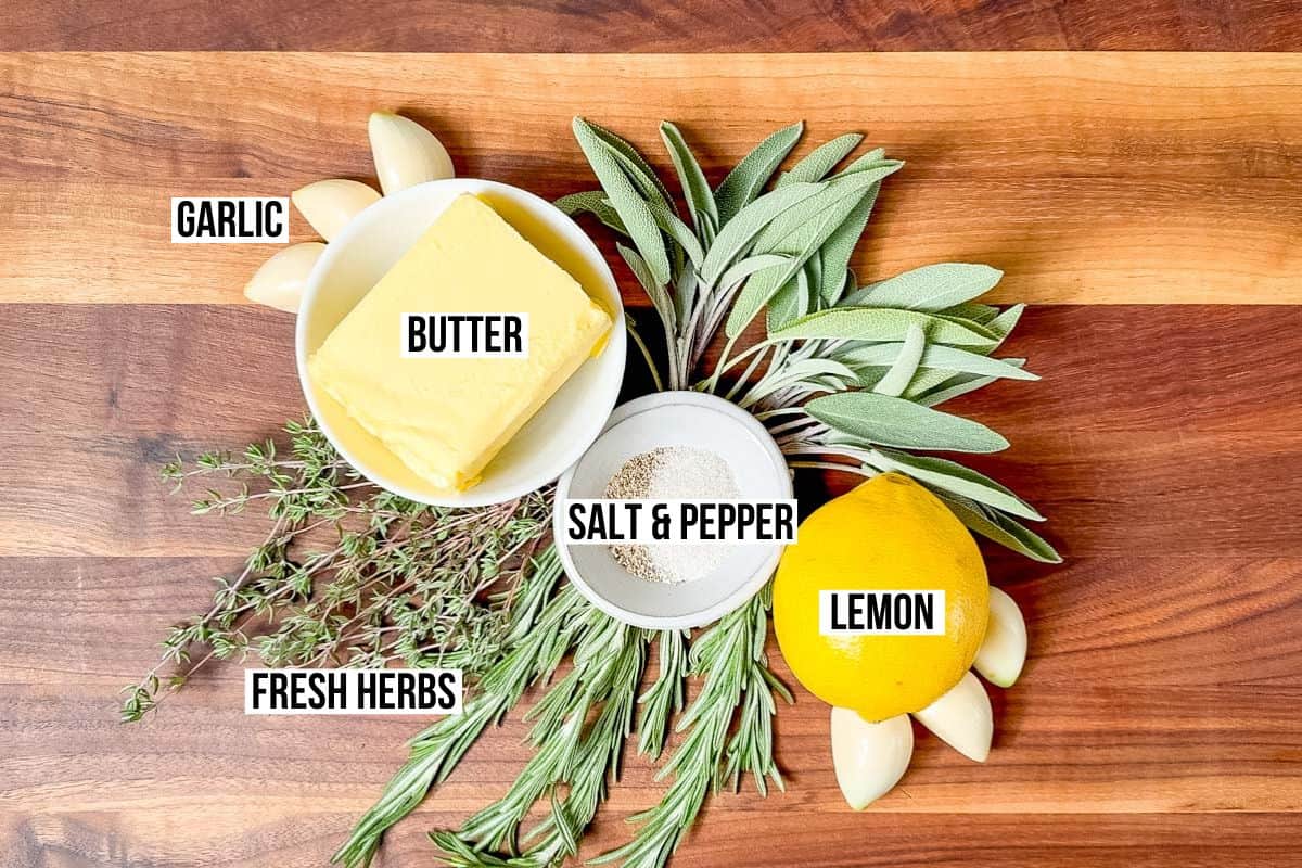 Butter, lemon, salt, pepper, garlic, rosemary, sage, and thyme on a wood cutting board.