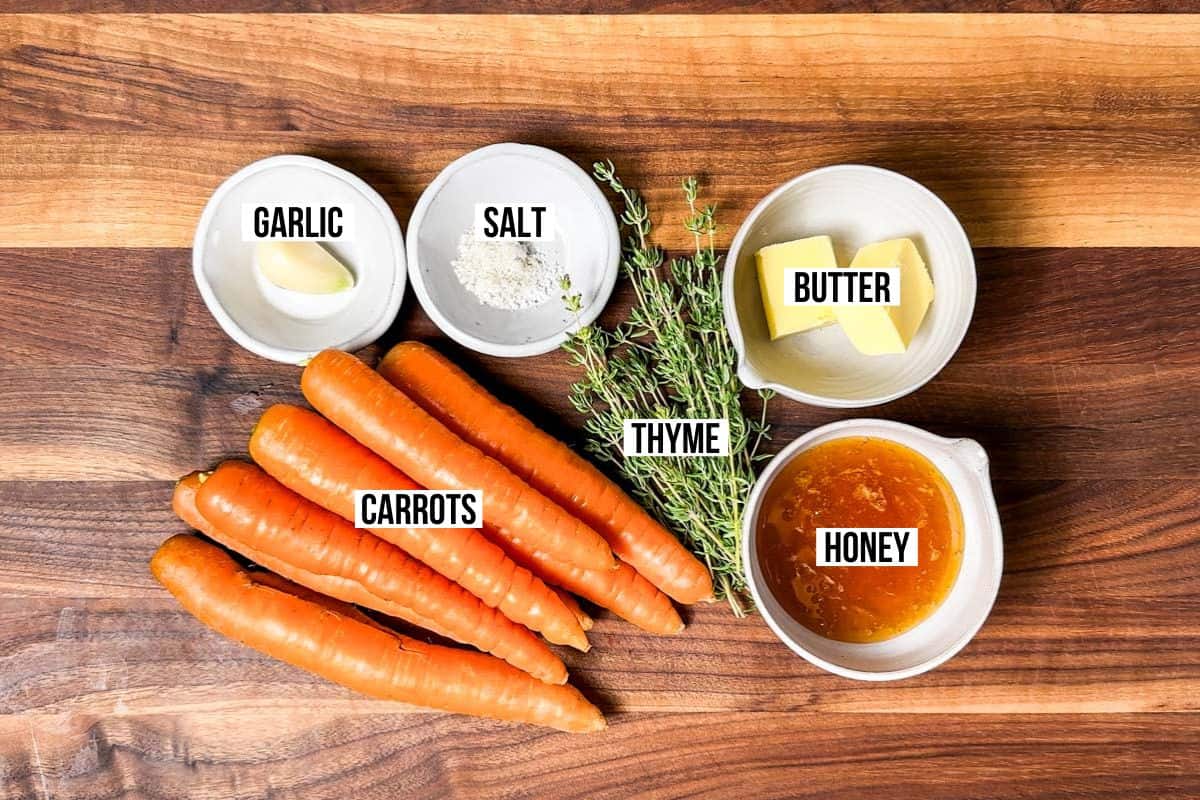 Carrots, garlic, thyme, honey, butter, and salt in bowls on a wood cutting board.