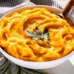 A white serving bowl filled with mashed butternut squash topped with a drizzle of butter and sage leaves with a wood spoon in it and sage leaves on the side of the bowl.