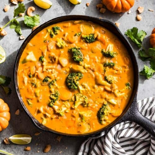 Finished pumpkin curry in a black cast iron skillet surrounded by lime wedges, cilantro, cashews, and mini pumpkins.