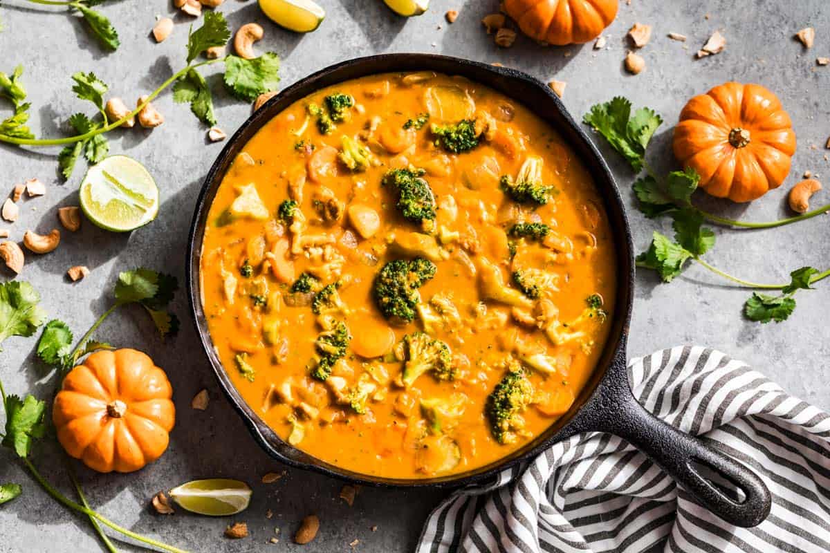 Finished pumpkin curry in a black cast iron skillet surrounded by lime wedges, cilantro, cashews, and mini pumpkins.
