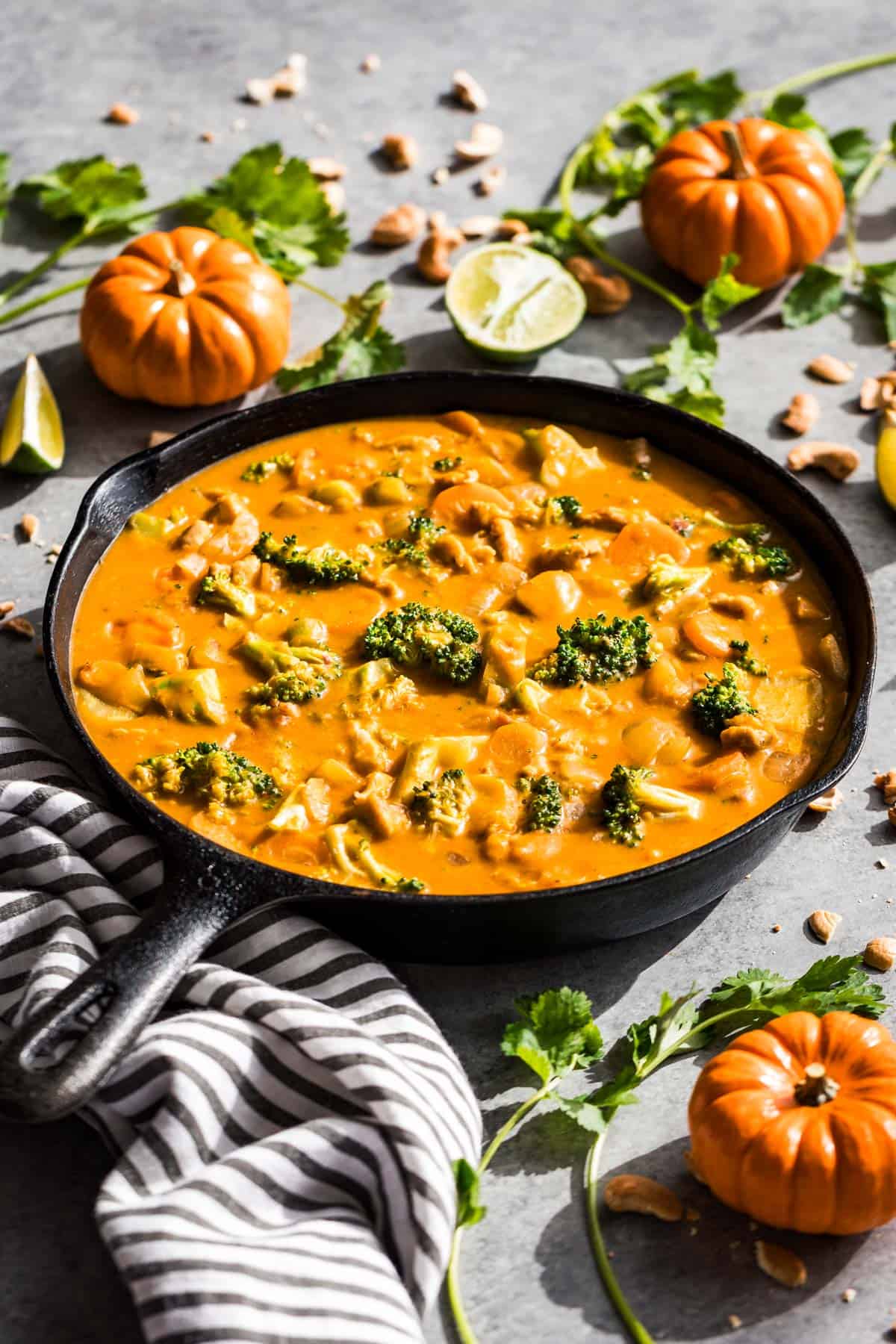 Thai pumpkin curry in a black cast iron skillet with mini pumpkins, cilantro, and lime wedges around it.