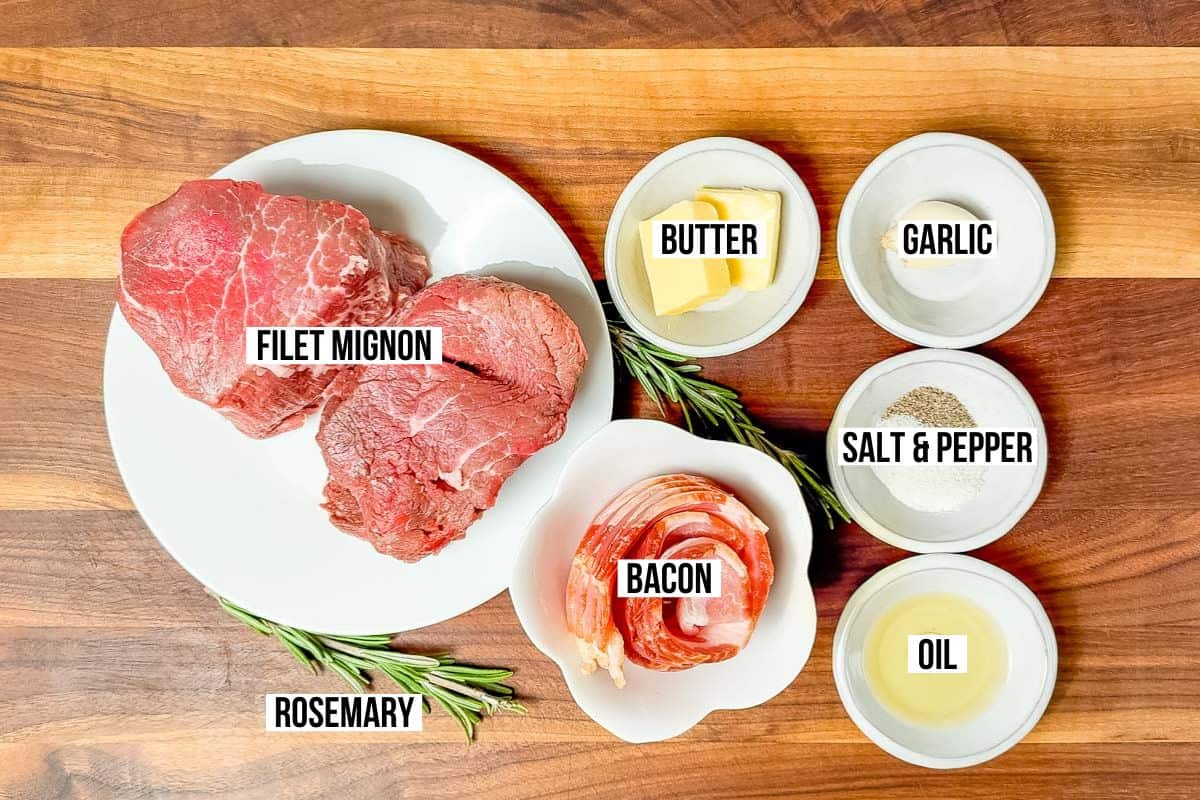 Two filet mignon steaks on a plate with bowls of bacon strips, oil, salt & pepper, garlic, butter, and rosemary on a wood cutting board.