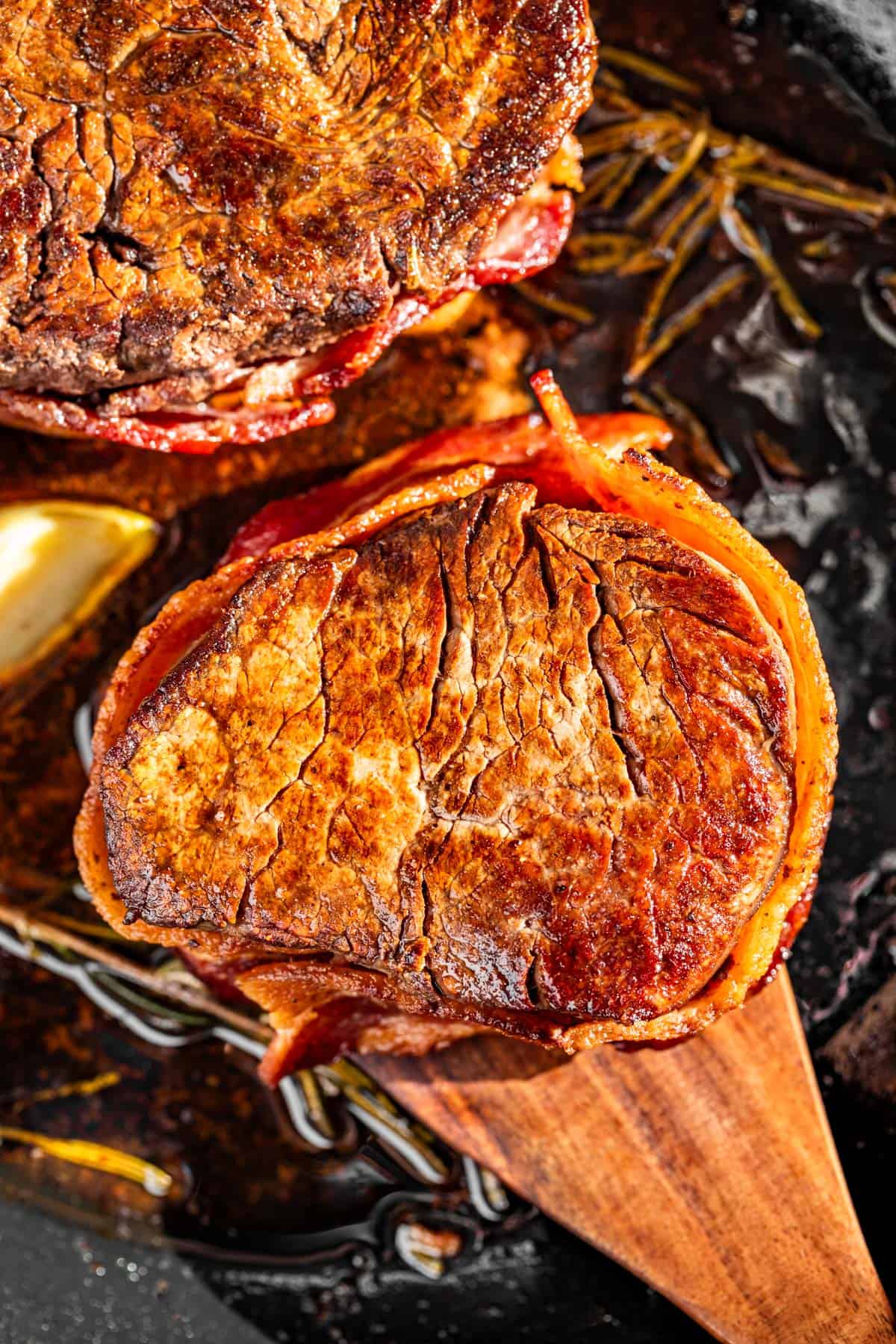 Two Bacon Wrapped Filet Mignons in a cast iron skillet with a wood spatula.