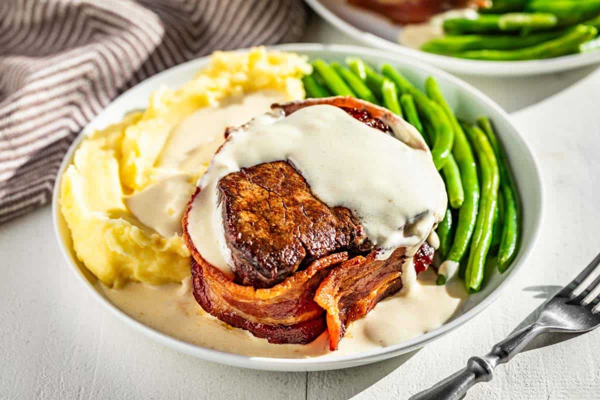 A bacon wrapped fillet topped with garlic cream sauce with a side of mashed potatoes and green beans on a plate.