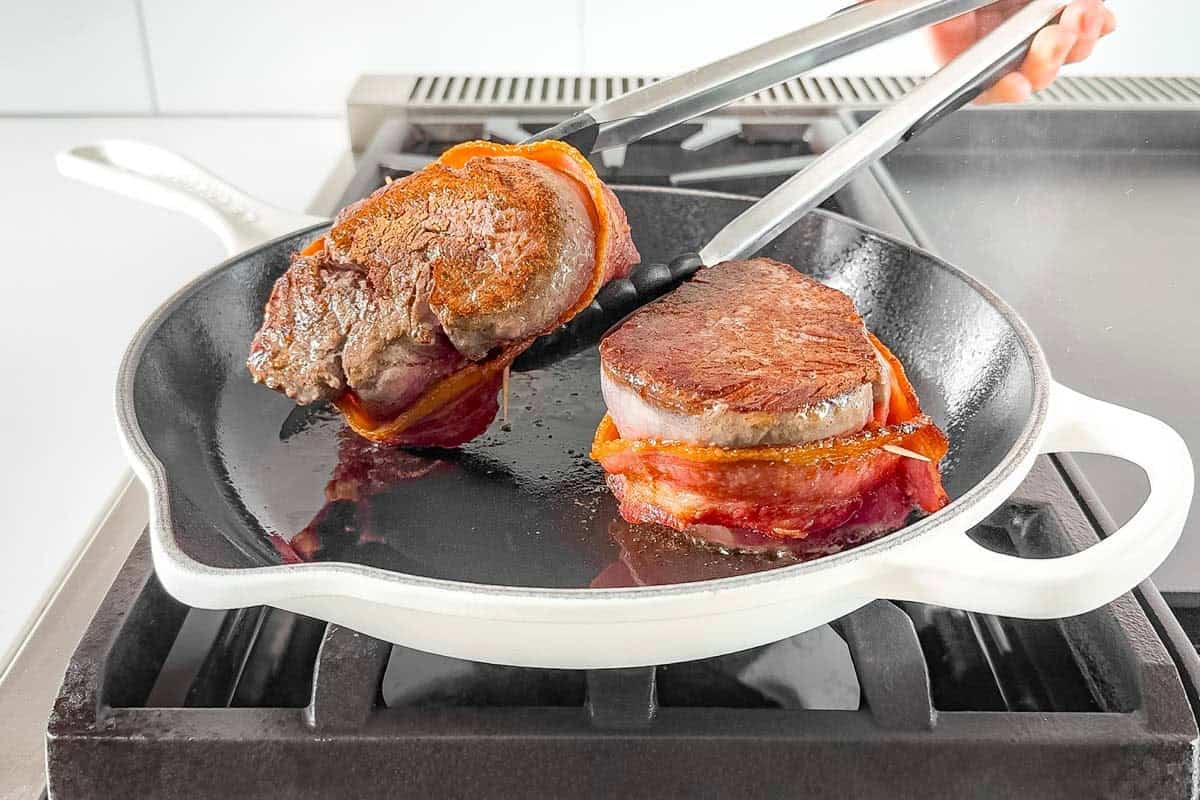 Searing the bacon wrapped steaks in a white cast iron skillet.