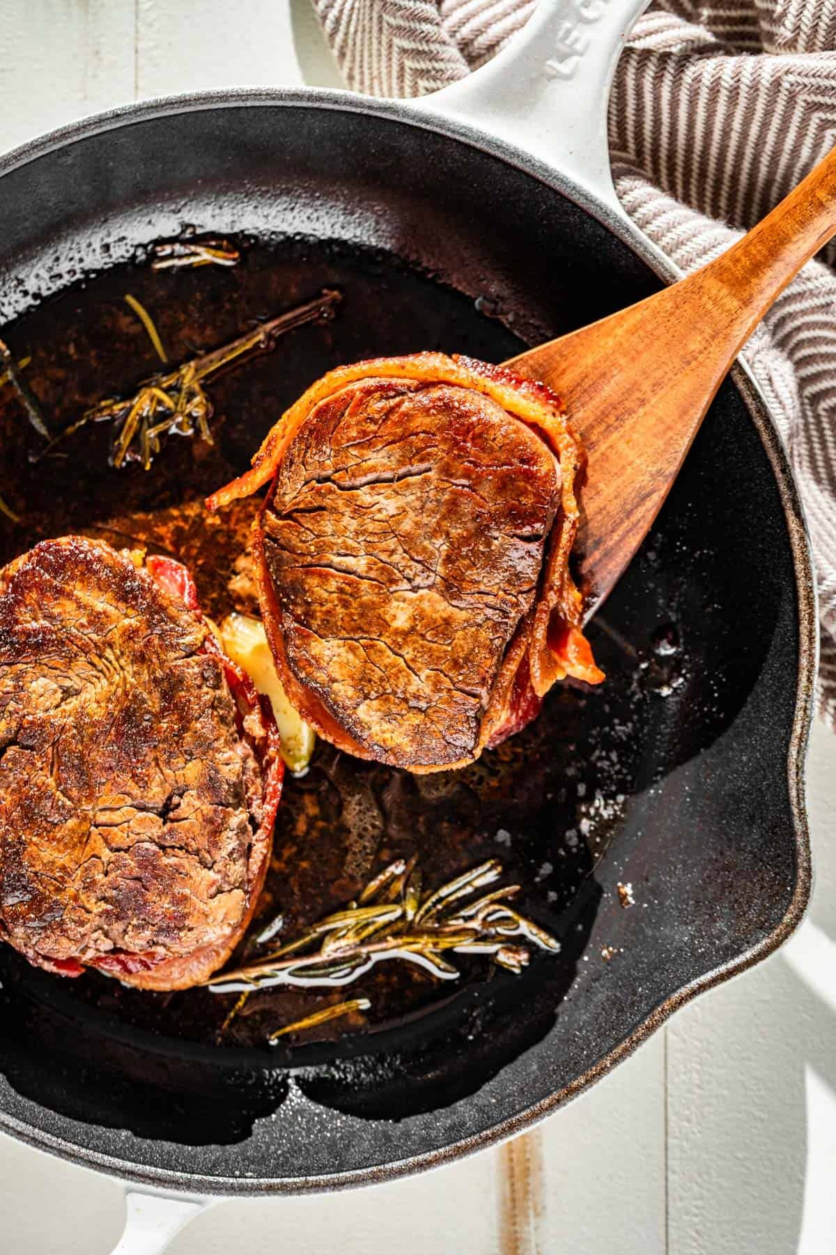 Two Bacon Wrapped Filet Mignons in a white cast iron skillet with a wood spatula scooping up one steak.