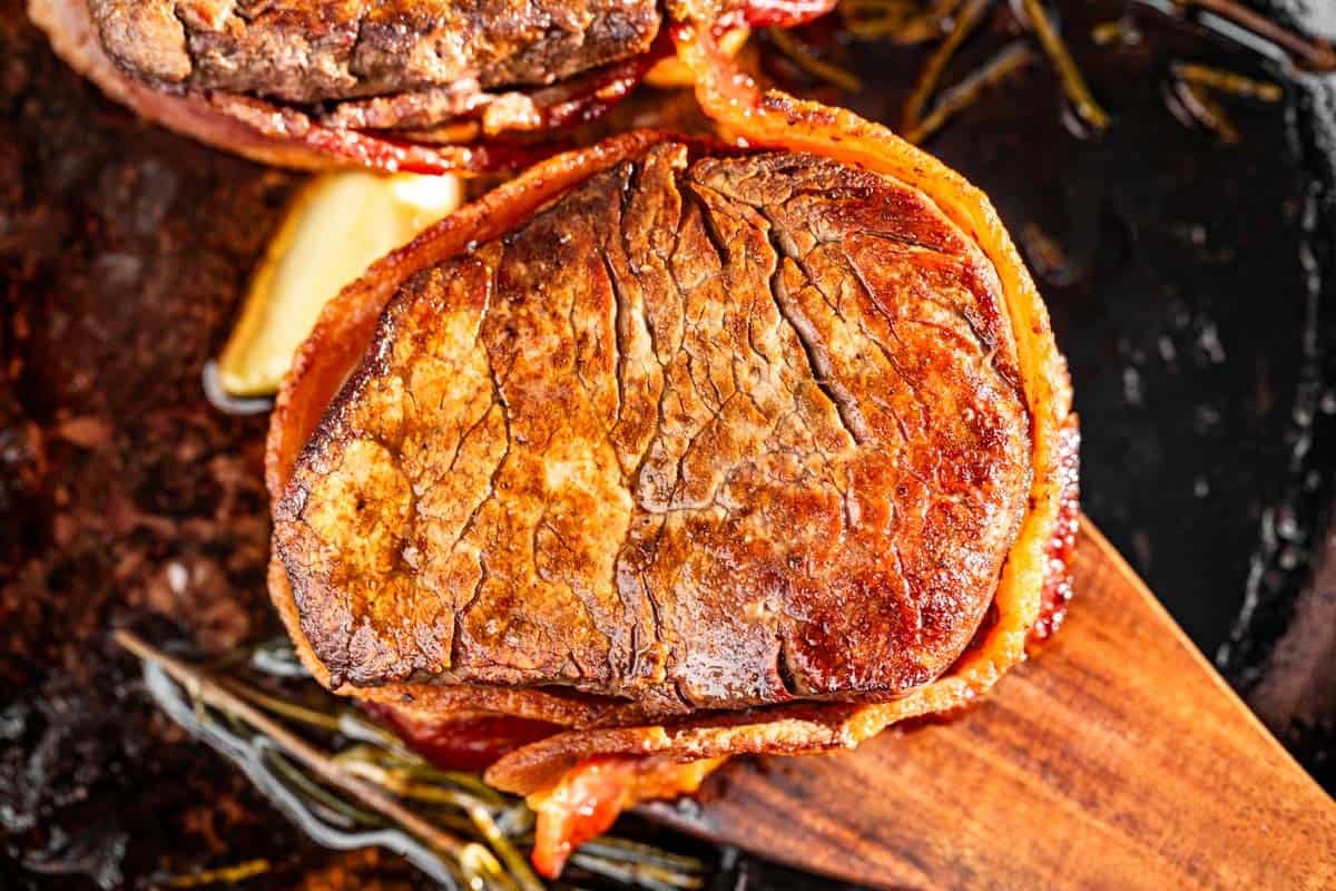 Two Bacon Wrapped Filet Mignons in a cast iron skillet with a wood spatula.