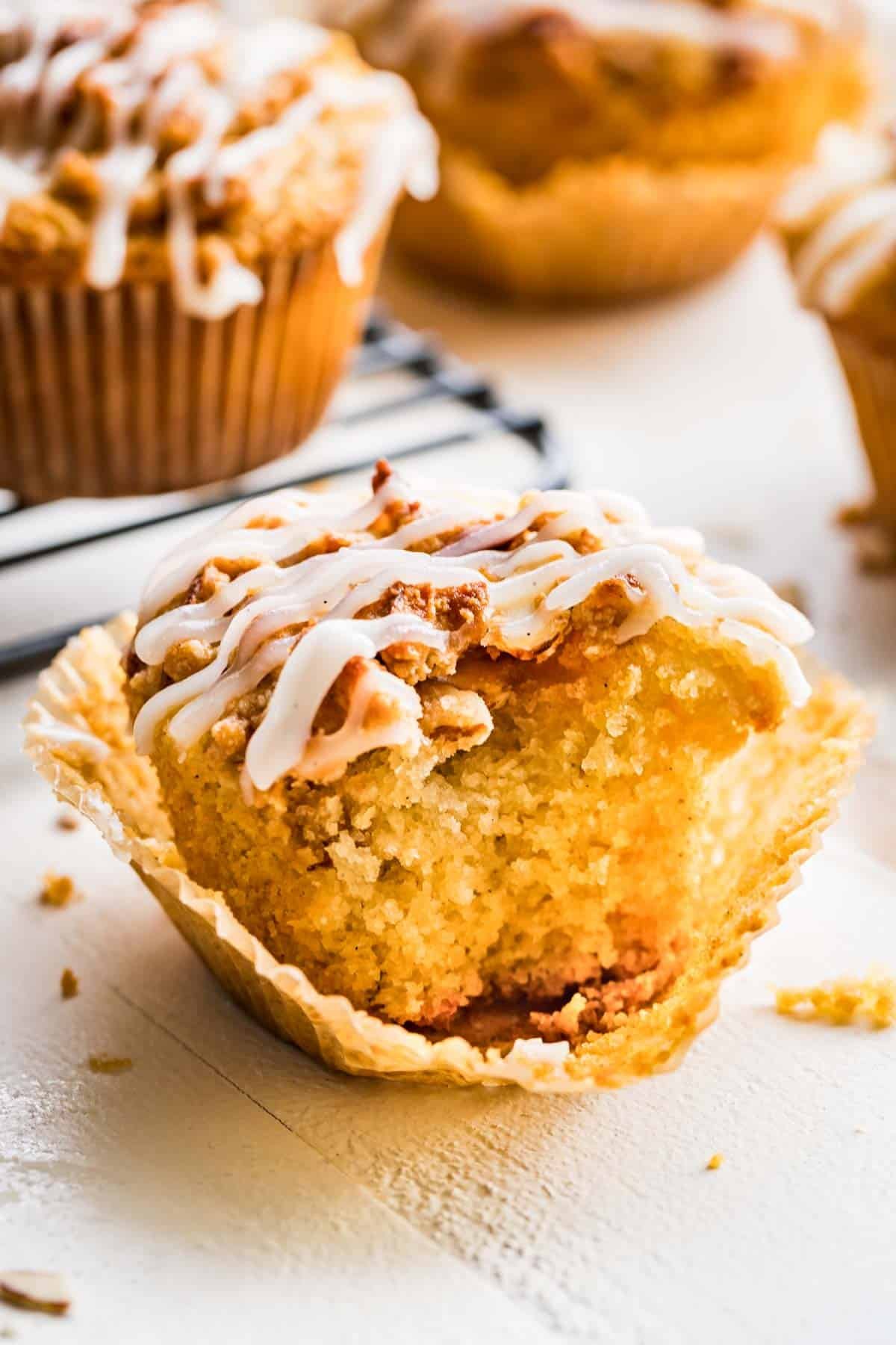 Vanilla almond flour muffins with streusel topping and a white glaze with the wrapper peeled back sitting on a white table.