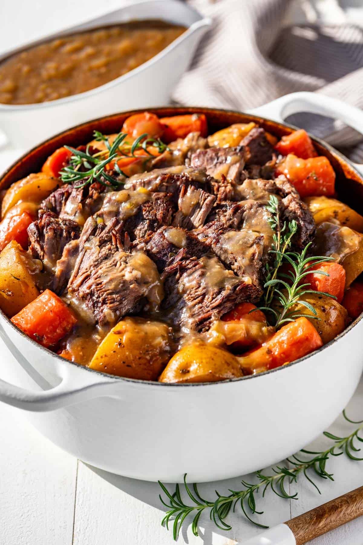 A large white Dutch oven filled with tender sliced pot roast, potatoes, and carrots with a gravy boat beside it.