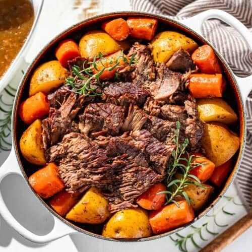 Finished sliced pot roast in a large white Dutch oven surrounded by potatoes and carrots and garnished with fresh rosemary.