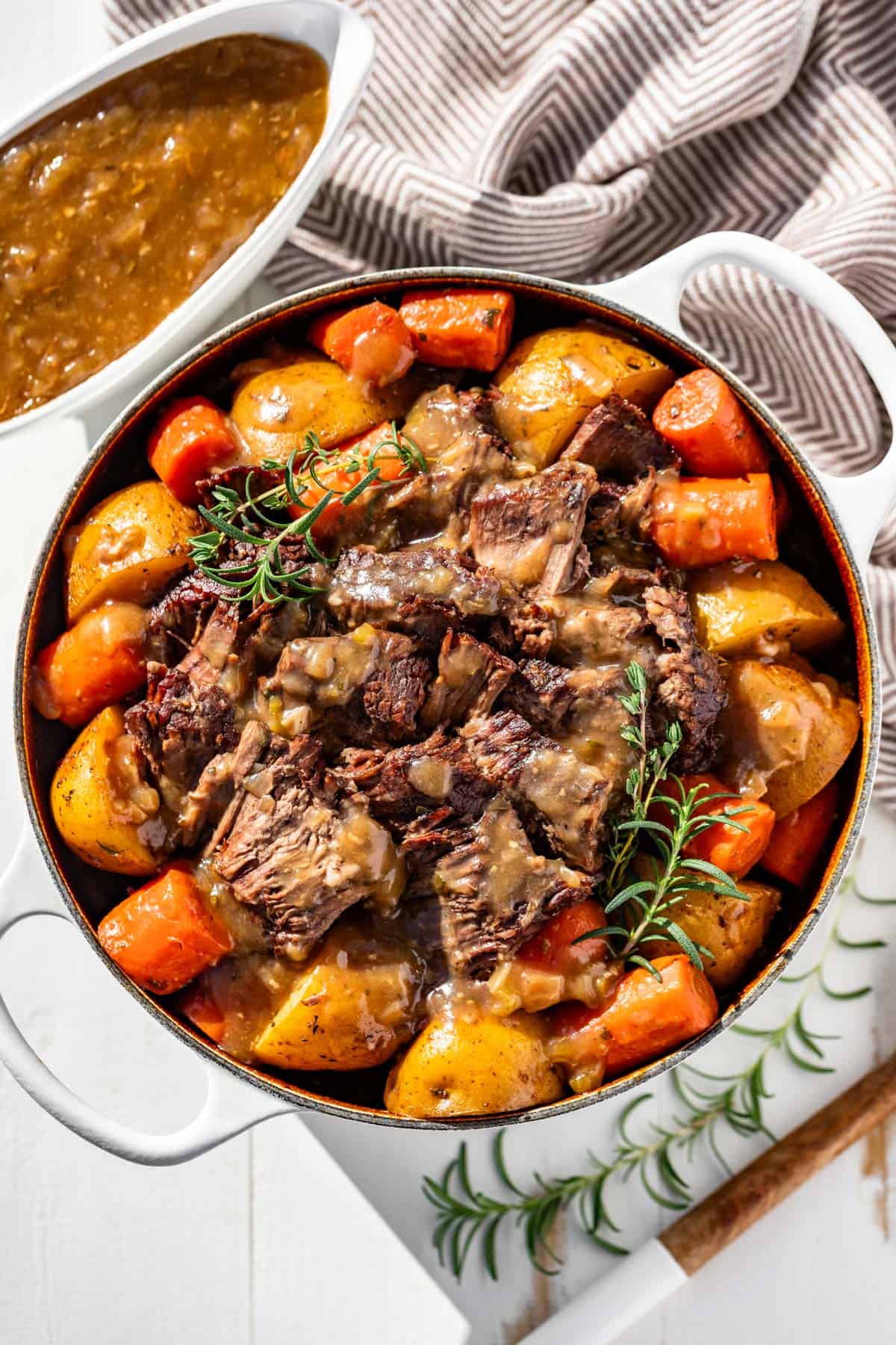A large white Dutch oven filled with roast beef, carrots, and potatoes, drizzled in gravy with a gravy boat full of gravy on the side.
