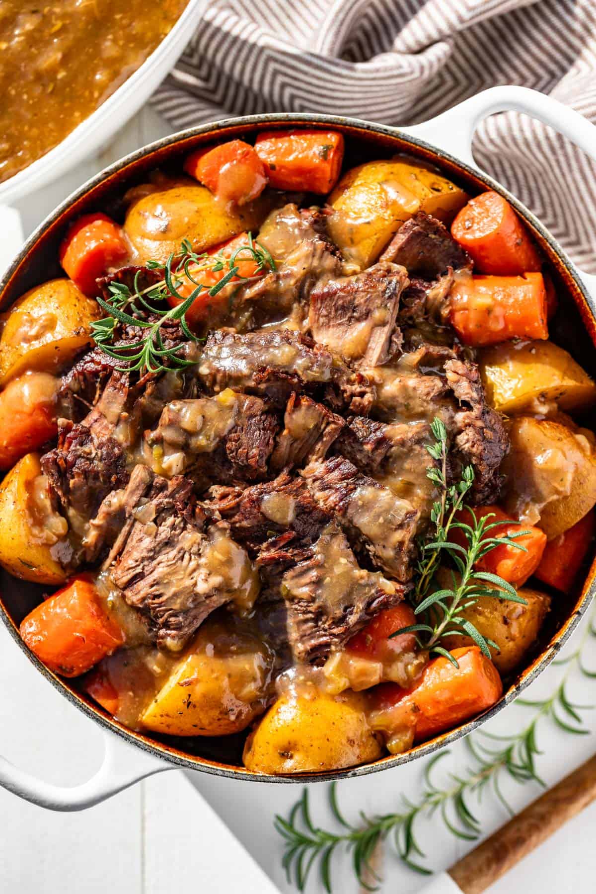 A large white Dutch oven with sliced pot roast surrounded by potatoes and carrots with a gravy boat full of gravy on the side.