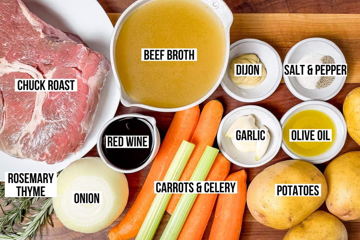 Chuck roast, beef broth, onion, rosemary, thyme, carrots, celery, red wine, garlic, potatoes, Dijon mustard, salt and pepper in small bowls on a wood cutting board.