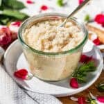 Sugar scrub in a glass jar with a gold spoon in it sitting on a white plate surrounded by roses and rose petals.