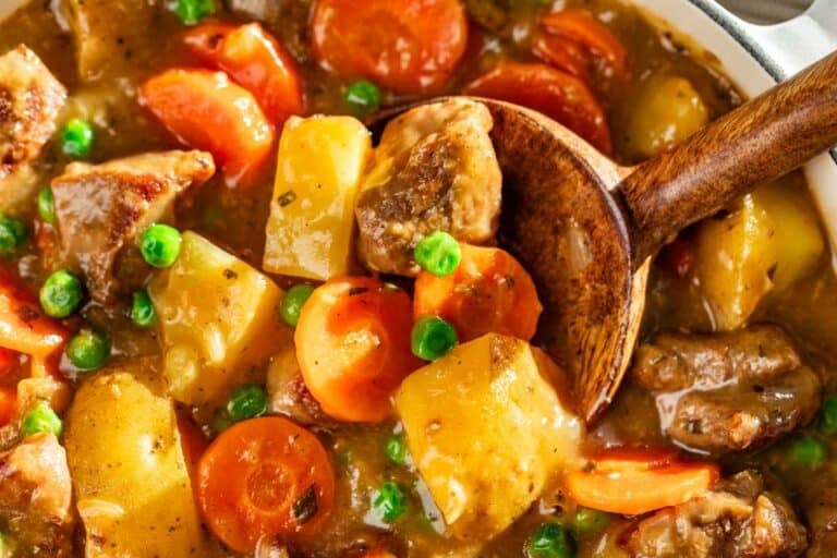 Very close view of a wood spoon scooping up pork stew with cubes of pork and potatoes, sliced carrots, and peas.