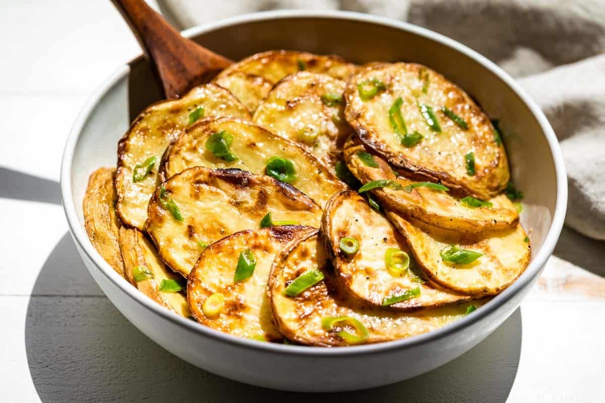 Sliced grilled potatoes sprinkled with green onions in a white bowl.