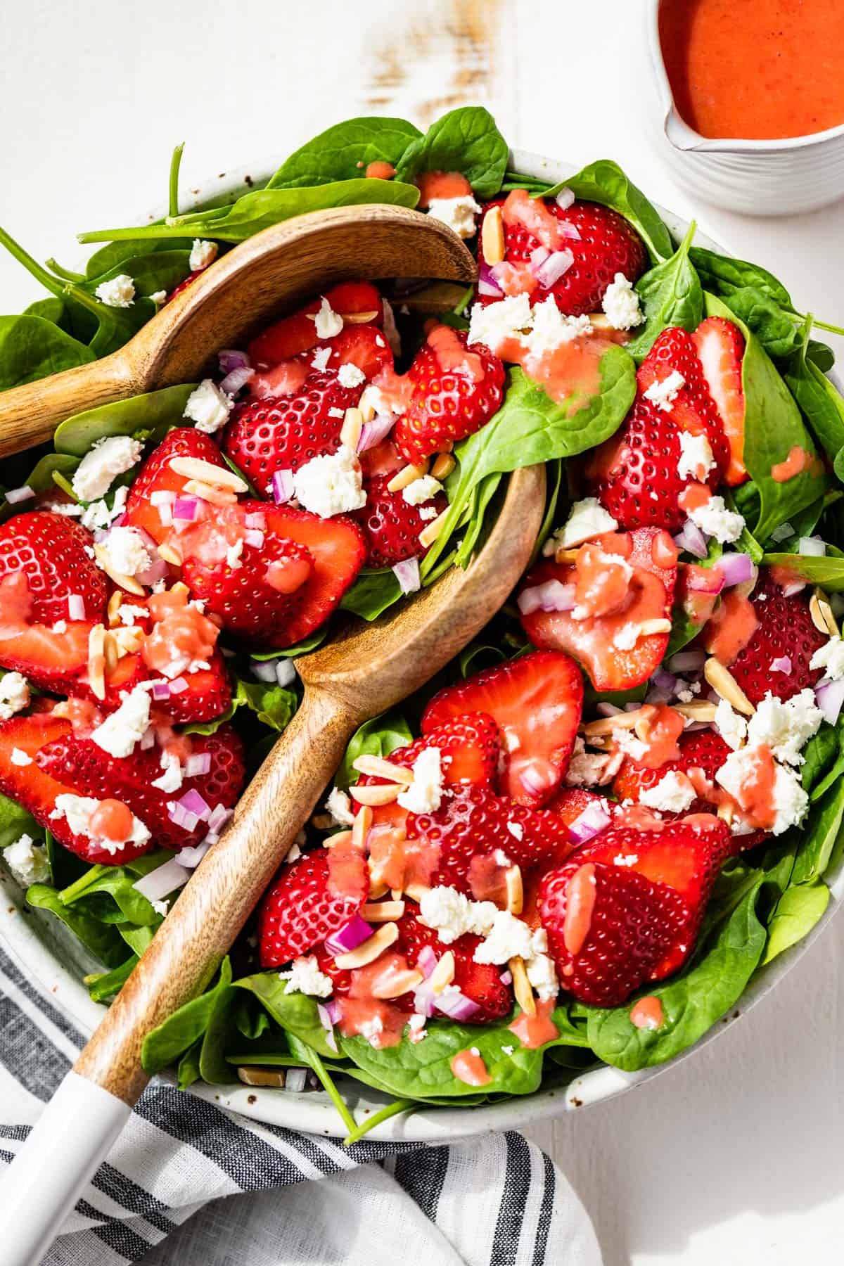 Strawberry spinach salad in a large white bowl with wood serving spoons and a side of strawberry vinaigrette.