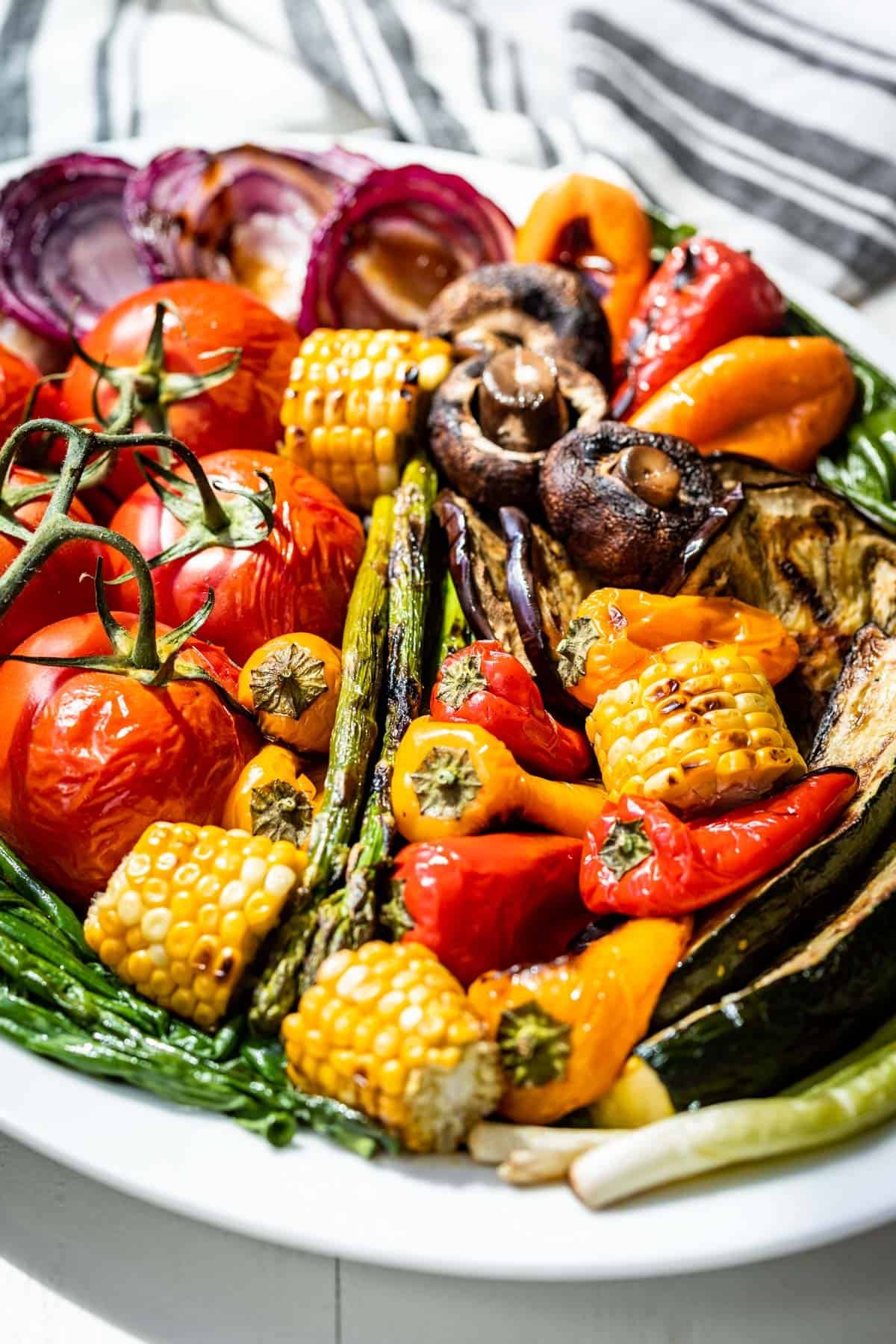 Grilled onions, corn, zucchini, peppers, asparagus, and tomatoes on a large white platter.