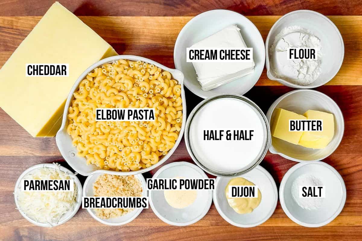 Cheddar cheese, elbow macaroni, parmesan cheese, cream cheese, Dijon mustard, garlic powder, butter, flour, and half and half in bowls on a wood cutting board.