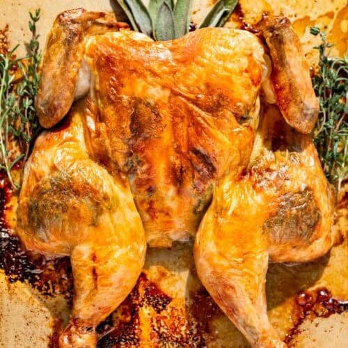 Finished Spatchcock Chicken on a gold baking sheet with rosemary, sage, and thyme sprigs around it.