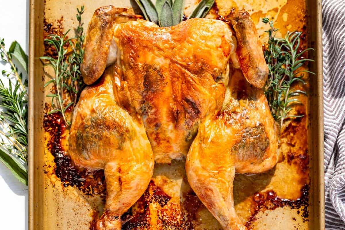 Finished Spatchcock Chicken on a gold baking sheet with rosemary, sage, and thyme sprigs around it.