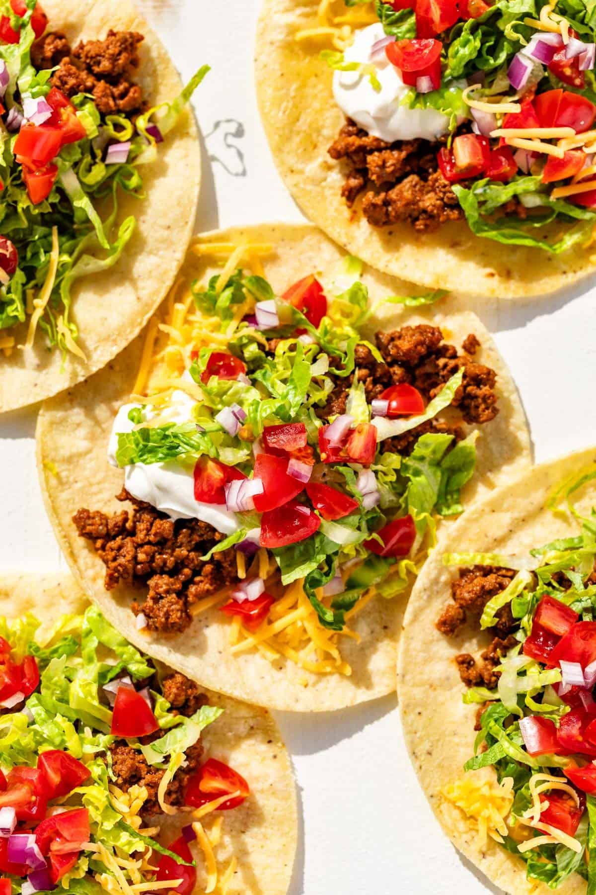 Five tacos with corn tortillas topped with taco meat, cheese, lettuce, tomatoes, and sour cream.