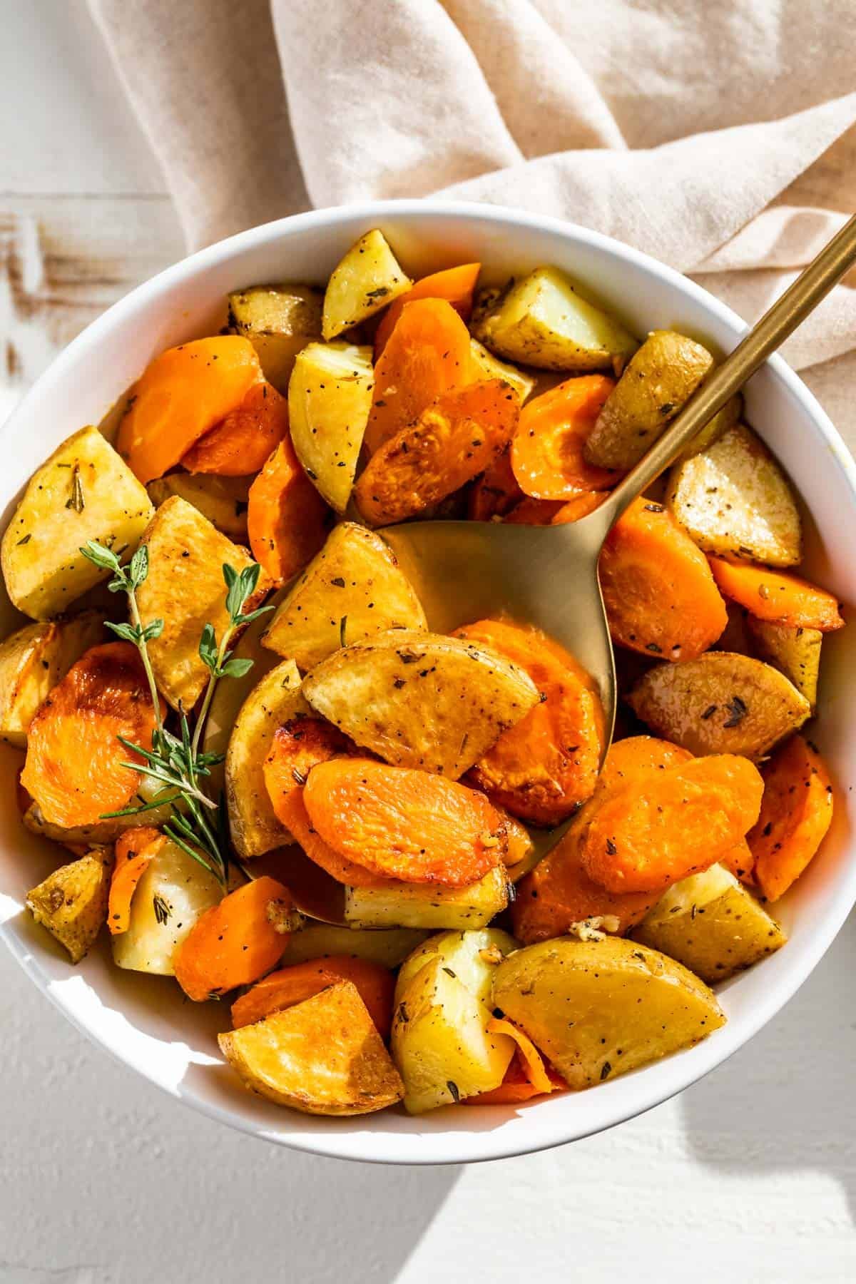 Roasted potatoes and carrots in a white bowl with a gold spoon scooping some out.